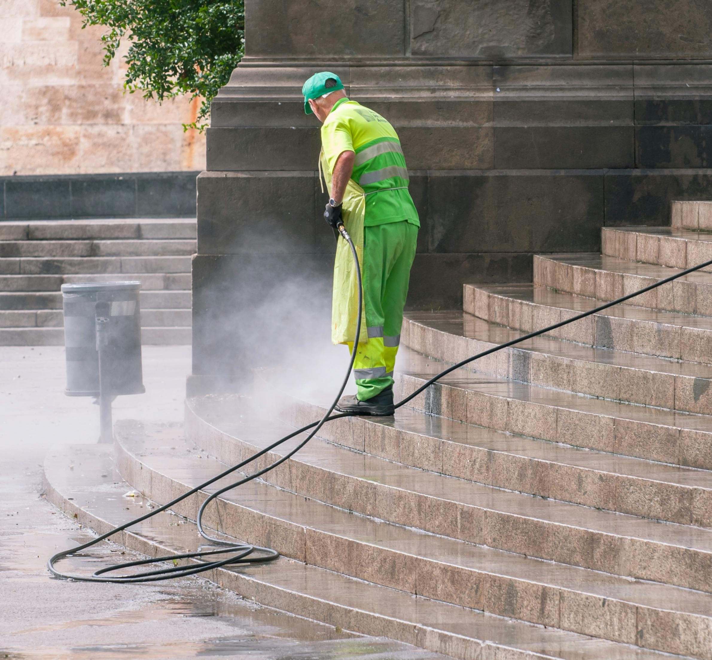 Worker in high-visibility gear pressure washing stone steps, creating a cloud of water mist.